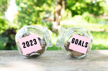 Glass jars with coins and the inscriptions 2023 and GOALS stand on a wooden table. Investment budget.