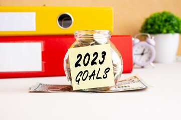 A glass jar full of coins sits on a stack of dollar bills on a work table. There is a sticker with the text 2023 GOALS.