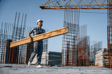 View from the side. Walking and holding wooden plank. Man is working on the construction site at...