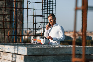 Standing and talking by phone. Businessman in formal clothes is on the construction site at daytime