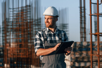 Looking to the side. Holding tablet. Man is working on the construction site at daytime