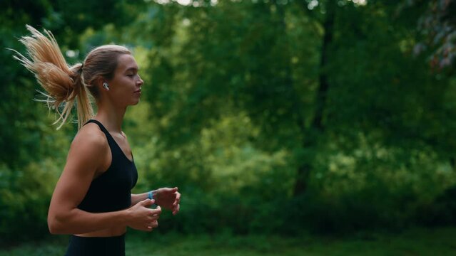 Side view of the female runner running wearing earphones in the park. Sportswoman morning jogging training. People, sport and technology concept.