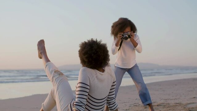 Lovely Black girl taking photo of mum at seashore while woman posing, sitting on sand. Mother and daughter having fun together on warm summer evening at seaside. Dolly shot. Childhood, hobby concept.