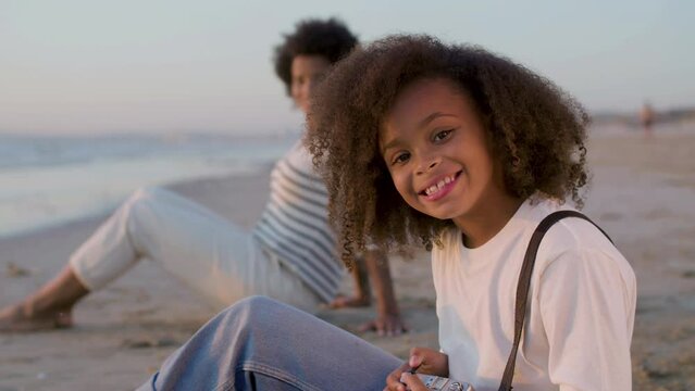 Closeup of cute Black girl spending time at beach with mum, sitting in background. Adorable child with thick curly hair holding camera, looking up and smiling happily at camera. Childhood concept.