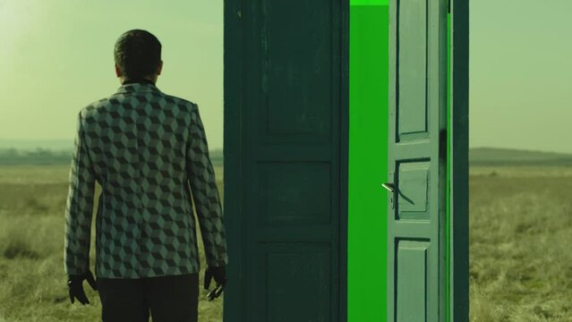Footage of man walking on field near door with green screen . Young stylish male artist standing on meadow field at day time . Concept of fantasy mystic dreams or teleportation. Another Dimension