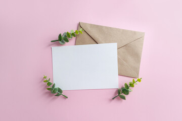 Blank sheet of paper and craft envelope on pink background surrounded by eucalyptus branches. Wedding mockup for invitation.