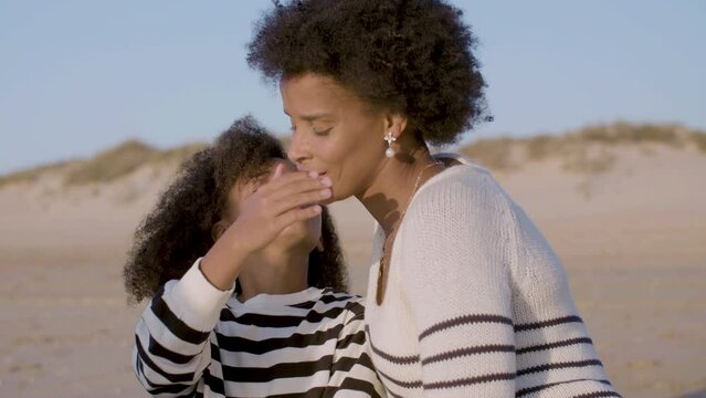 Cute girl kissing her mum on cheek, spending time on beach together. Beautiful smiling Black woman and her daughter enjoying sunset at seashore. Handheld, medium closeup shot. Happy family concept.