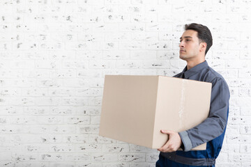 Handsome man from moving service carrying heavy box in front of white brick wall background	