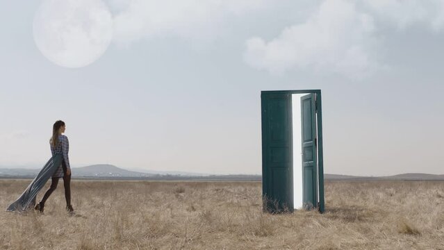 Footage of female model walking on field near door . Young stylish lonely girl stand on meadow field at day time . Long skirt fabric flowing by wind . Concept of fantasy mystic dreams or teleportation