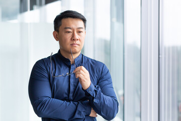 Portrait of a serious young Asian businessman, lawyer. He stands thoughtfully in the office by the window, holds glasses in his hands, looks to the side.