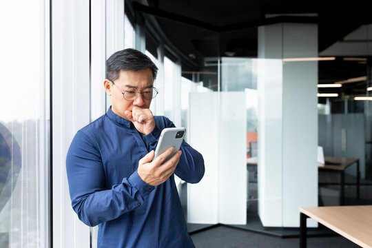 A Worried And Serious Young Asian Man Is Standing In The Lobby Of An Office, Hospital, Bank. Holds The Phone In His Hands, Looks At The Screen In Shock, Received And Reads Bad News, Dials A Message.