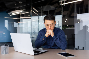 Serious male Asian businessman sits in the office at the table, works with a laptop and documents. He holds his head in his hands, thinks, solves a problem.