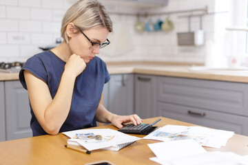 A woman with glasses at the table counts money and looks at bills for communal services. The girl counts the family budget on a calculator and keeps a note in a notebook.