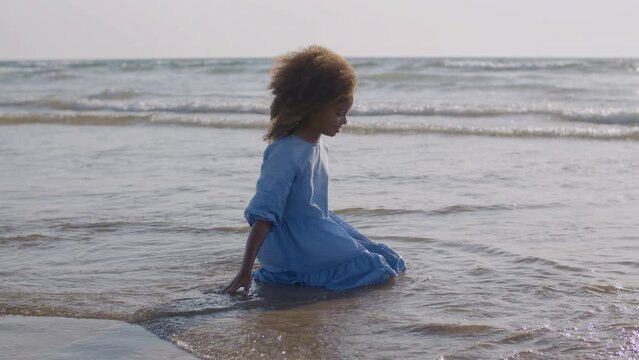Cute Black girl sitting kneeling on seashore with waves around rushing to shore. Happy child in blue dress smiling and touching water while wind playing with her hair. Happy childhood, holiday concept