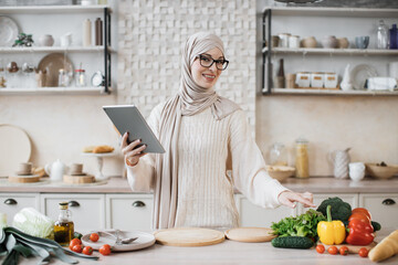 Happy muslim woman in hijab cooking healthy food pointing on vegetables lying on counter while reading recipe on digital tablet in modern kitchen at home. Preparing vegetable salad.
