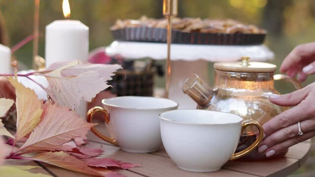 Cozy autumn picnic in the park. Close up of unrecognisable woman pouring hot tea from glass kettle into classic cups. Homemade apple pie, maple leaves and burning candles on wooden table outdoors