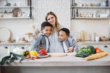 Healthy food, home resting concept. Pretty young mother and two children sons preparing salad with fresh vegetables, hugging and smiling looking at the camera while cooking in kitchen.