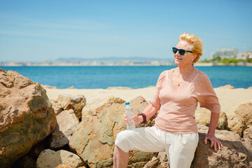 Retro style portrait of attractive senior woman sitting on the beach with a bottle of water wearing dark sunglasses. Old woman enjoying life