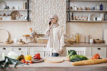 Fototapeta na wymiar Portrait of young beautiful muslim woman in hijab standing at bright modern kitchen, using phone during call with her friends while cooking vegan salad from fresh vegetables at home.