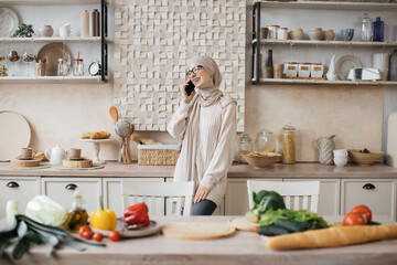 Portrait of young beautiful muslim woman in hijab standing at bright modern kitchen, using phone during call with her friends while cooking vegan salad from fresh vegetables at home.