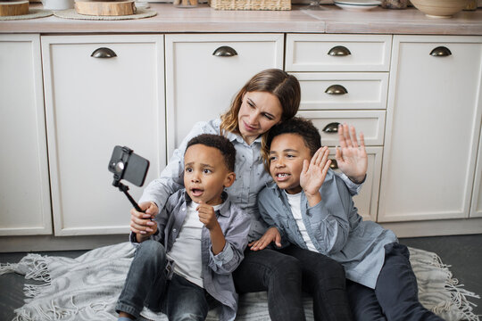 Happy multinational family of mother and two sons sitting on warm floor in light kitchen during weekend, resting and taking positive photo using selfie stick and waving hands.