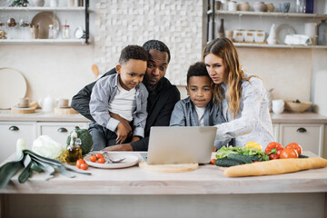 Excited african little boys with their father and caucasian mother reading healthy salad recipe on laptop, sitting at wooden table in kitchen, multiracial family having fun, enjoying weekend.