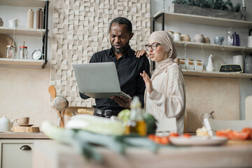 Young wife and husband looking at online recipe on a digital laptop pc. Attractive muslim female and african male surfing the internet for recipes while making vegetarian salad in bright kitchen.