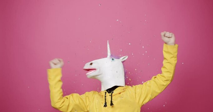 Man in unicorn mask dancing in confetti. Happy guy wearing mask of unicorn and yellow hoodie shaking raised arms while standing under falling glitter confetti on pink background