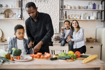 Selective focus on loving mom and little boy child having fun together in the kitchen. Blur view of african father teaching another little preschooler son to chop vegetables preparing salad for lunch
