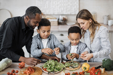 Close up view of parents and their sons cooking in kitchen preparing food salad together. Happy boys children assist mom and dad, helping with dinner or lunch, tearing leaves of lettuce into bowl.