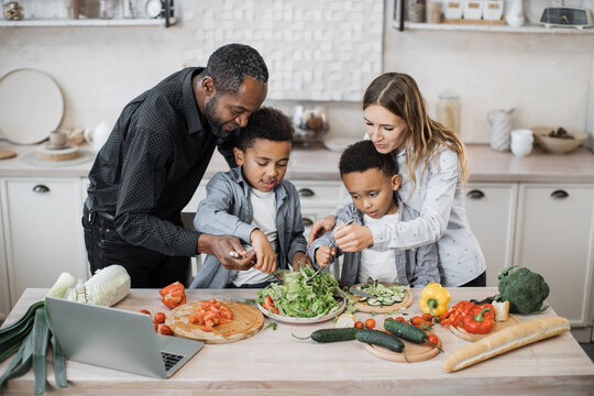 Young Parents Teaching African Little Cute Sons Preparing Healthy Vegetarian Meal With Sliced Vegetables. Mom And Dad Sharing Salad Recipe, Multiracial Family Chatting Enjoying Time In Kitchen.