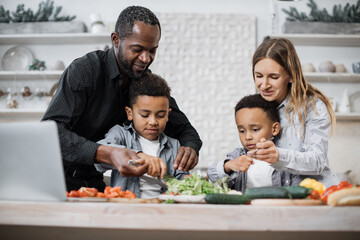 Young parents teaching african little cute sons preparing healthy vegetarian meal with sliced vegetables. Mom and dad sharing salad recipe, multiracial family chatting enjoying time in kitchen.