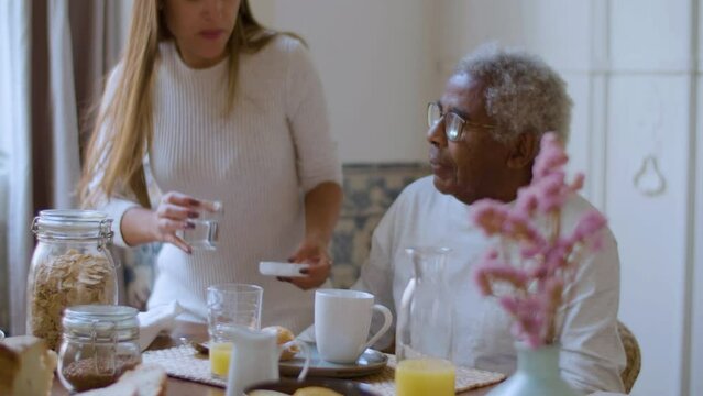 Black elderly man having breakfast when woman bringing him pills, reminding of taking medication. Pretty Caucasian lady taking care of senior relative. Relationship, elderly care concept.