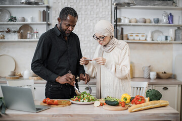 African american man holding spoon and fork, mixing ingredients in bowl, preparing delicious healthy salad, meal on table dinner in kitchen, next to his wife taking photos for social media.
