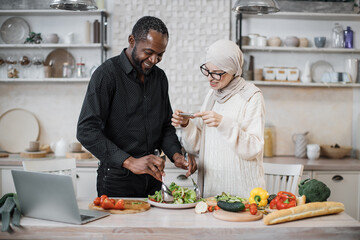 African american man holding spoon and fork, mixing ingredients in bowl, preparing delicious healthy salad, meal on table dinner in kitchen, next to his wife taking photos for social media.