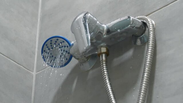 Shower head in bathroom with low water pressure. Chrome faucet and grey tiles on the walls, water flows towards camera, weak water pressure