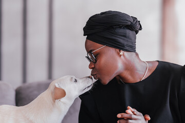 Close up shoot of Young African woman in black traditional clothes kissing her  white dog at home....