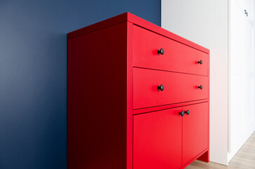New red chest of drawers in the interior of the apartment against the background of a blue wall and a white wardrobe