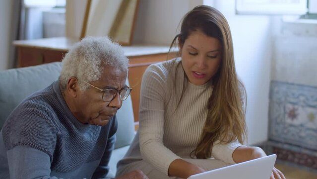 Pretty Caucasian girl helping senior Black man with using laptop. Young lady sitting on couch with her grandpa while explaining things and pointing at screen. Elderly care, generations concept.