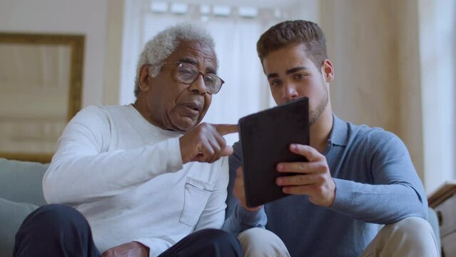 Bearded Caucasian guy helping senior Black man with using tablet. Young male sitting on couch with his grandpa while explaining things and pointing at gadget screen. Technology, generations concept.