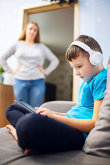 Mother scolds her son. Boy uses tablet with headphones and ignores his mom