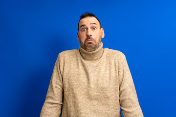 Fototapeta premium Frontal portrait of a hispanic man with a beard wearing a brown turtleneck doubting shrugging isolated on a blue background.
