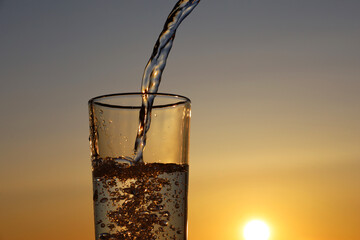 Clean water pouring into drinking glass on sunset sky background. Concept of health and freshness,...