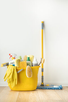 Brushes, Bottles With Cleaning Liquids, Sponges, Rag And Yellow Rubber Gloves On White Background. Cleaning Supplies In The Yellow Bucket On The Wooden Floor. Cleaning Company Service Advertisement