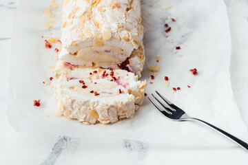 Cut of homemade meringue roll decorated with almond flakes and freeze-dried red berries on the white marble background. Meringue roulade with whipped cream on a parchment paper
