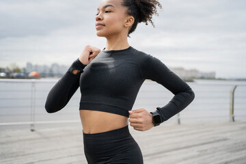 The fast runner trainer is focused on the routine work on the body. On the arm uses a fitness watch for sports. A young adult woman trains and runs in comfortable sportswear.