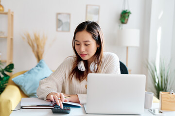 Concentrated young female remote worker in casual clothes sitting at table with laptop and taking notes in notebook while working