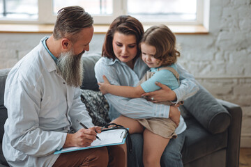 Kind male senior paediatrician doctor visiting his patient at home, examining little girl sitting on mother's lap, writing prescription. Concept of kid's health check