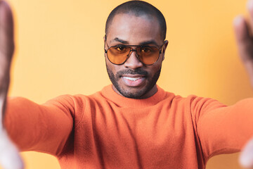 Obraz premium Smiling dark-skinned young man in sunglasses taking a photo, selfie self-portrait with mobile phone while holding the cellphone with two hands on an orange wall.