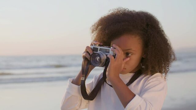 Cute Black girl spending time at seashore, taking photos in early evening. Pretty young girl with thick curly hair making shot with digital camera. Static shot. Childhood, hobby, leisure concept.